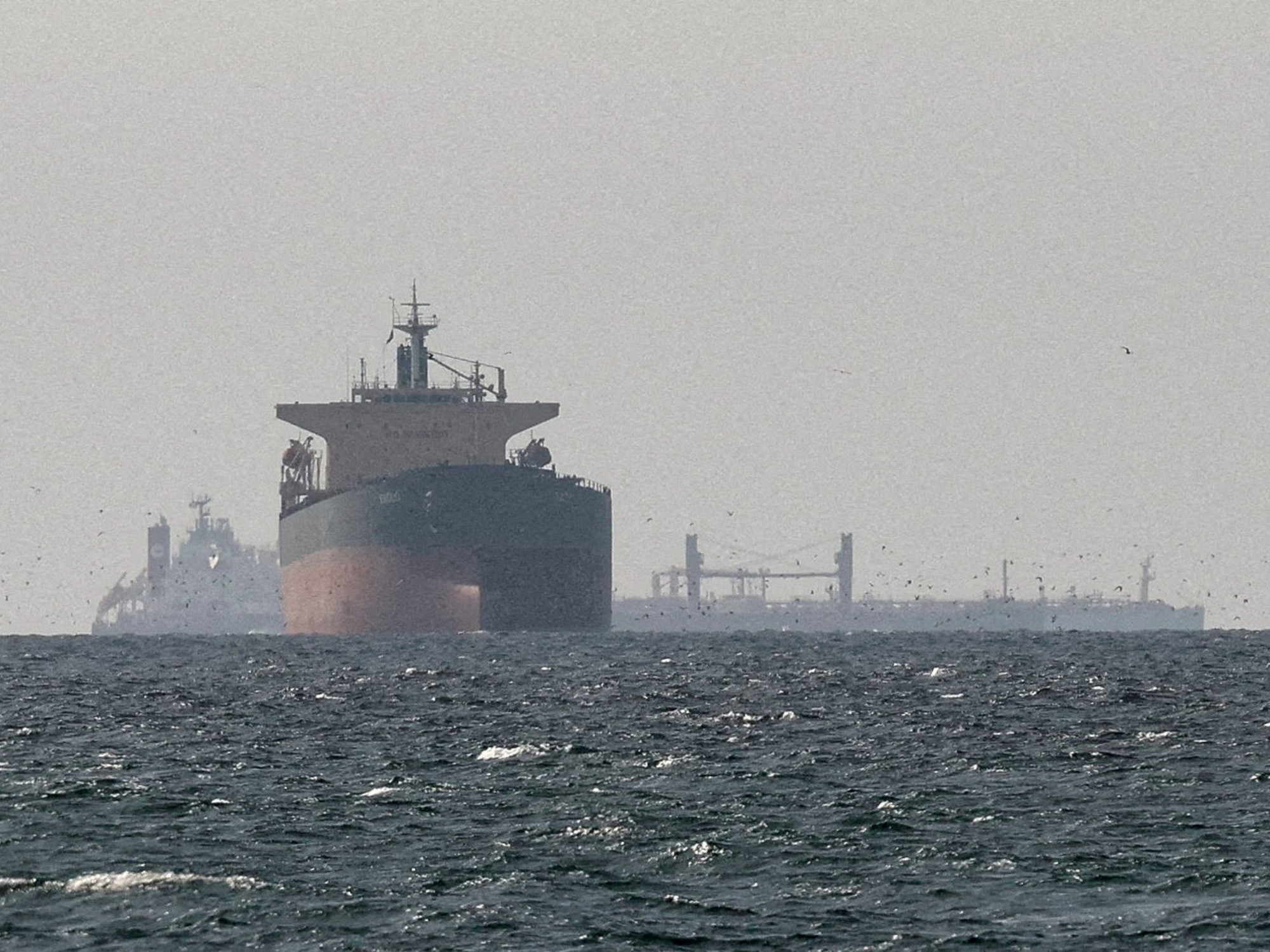 Un buque carguero navegando por el Estrecho de Ormuz, con grandes tanques de almacenamiento de petróleo visibles y la costa montañosa al fondo.