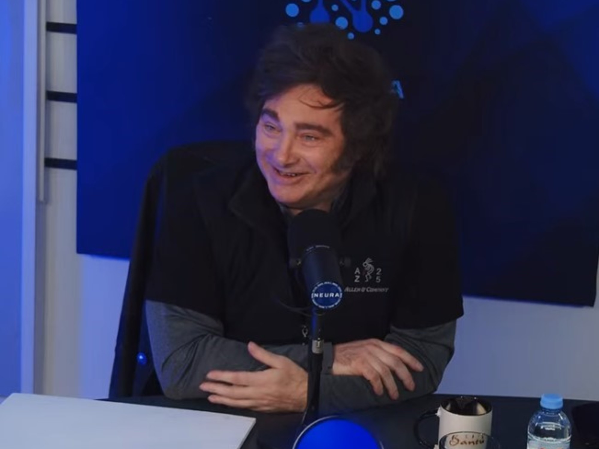 Javier Milei durante una entrevista en streaming, con elementos visuales que sugieren actividad en redes sociales.