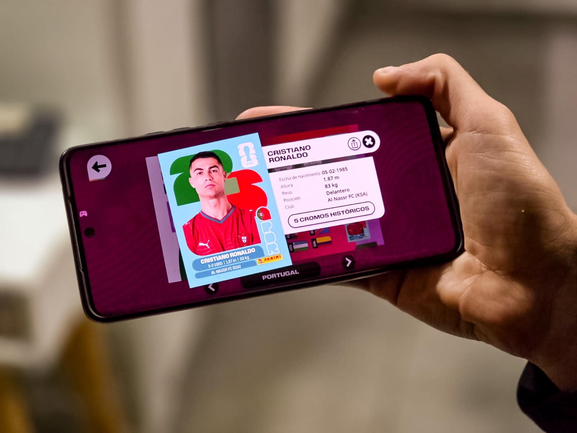 Un joven sonríe mientras interactúa con la aplicación del álbum digital del Mundial 2026 de Panini en su teléfono móvil, con el logo de Panini visible en la pantalla.