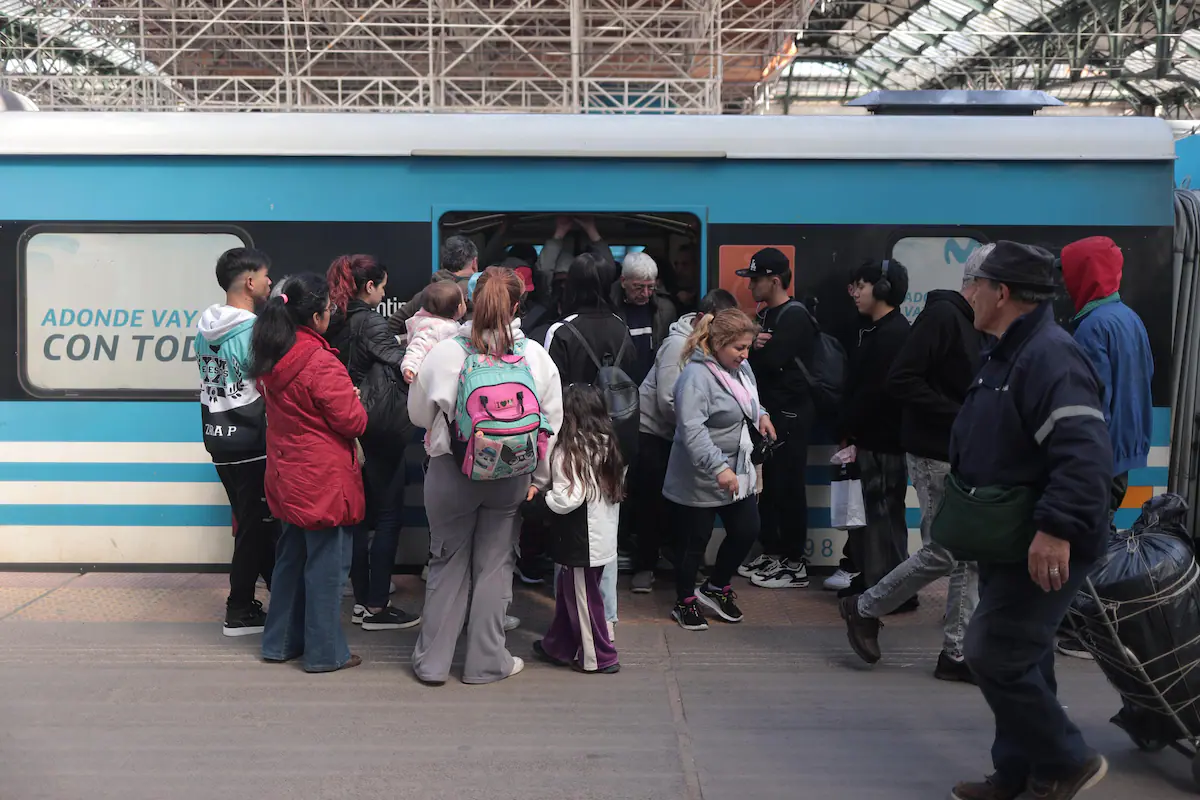 Un tren de pasajeros en una estación de Buenos Aires, con colectivos circulando en segundo plano, ilustrando el transporte público en el AMBA.