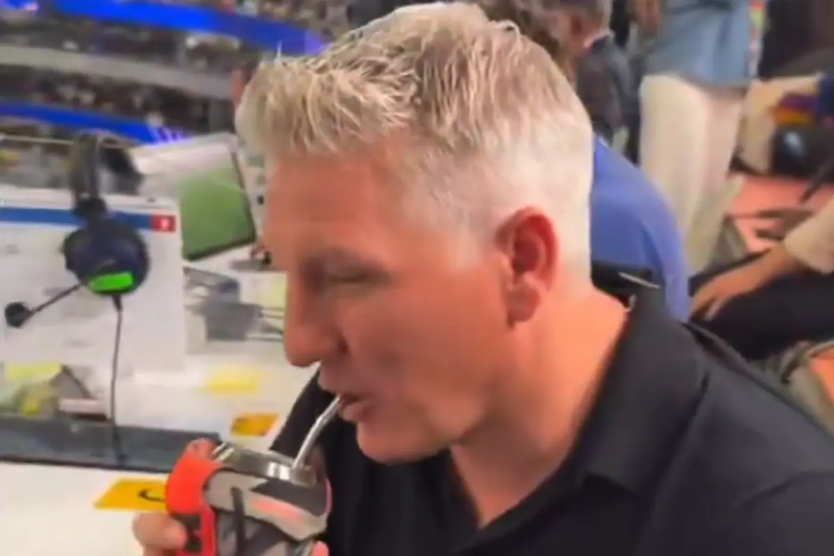 El exfutbolista alemán Bastian Schweinsteiger sonríe mientras sostiene un mate y bebe de él, con un fondo de estadio deportivo.