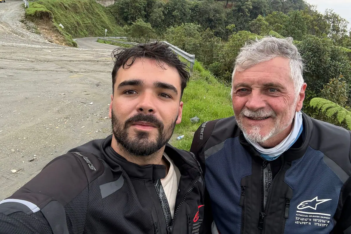 Padre e hijo sonriendo junto a sus motocicletas en una ruta escénica.
