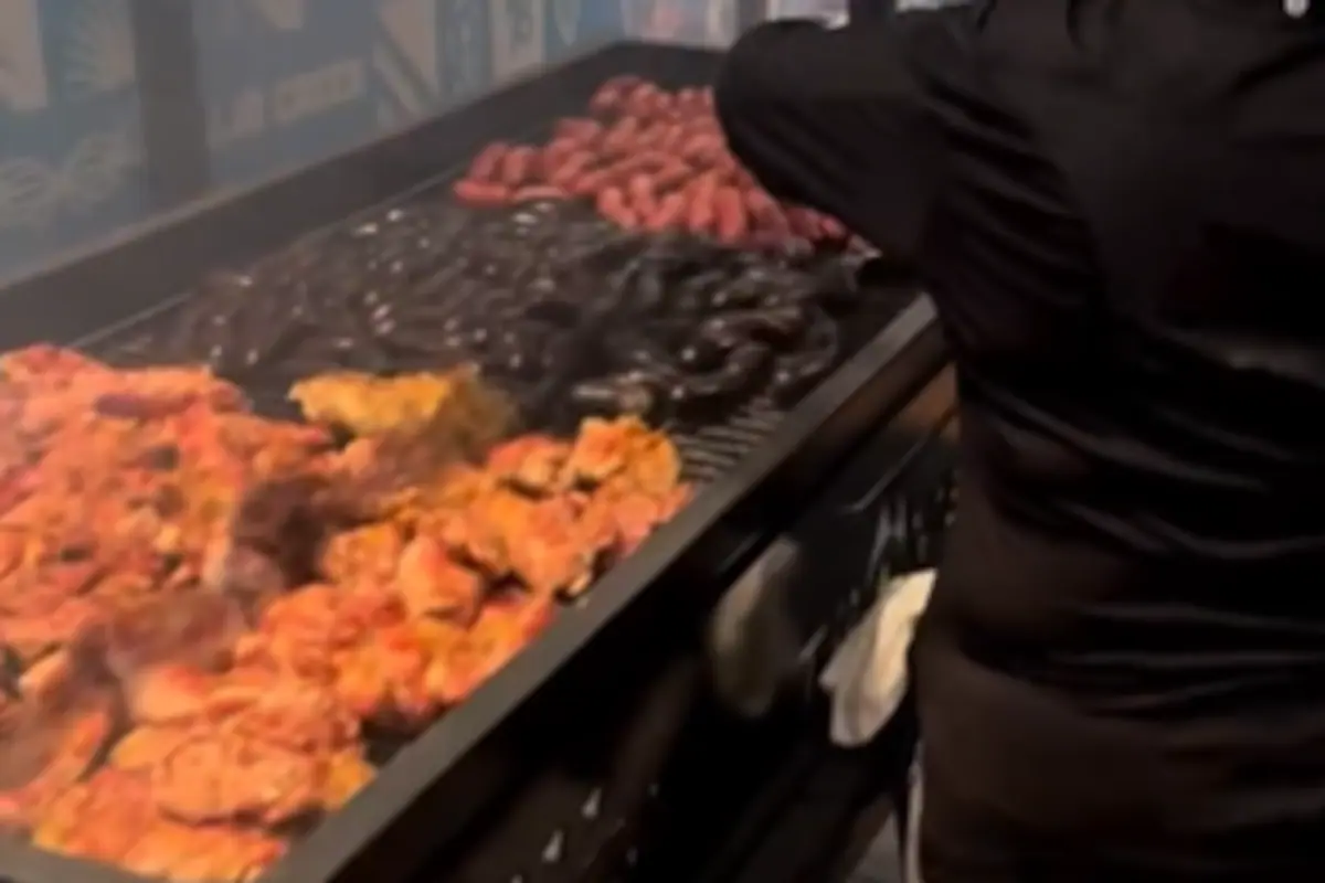 Emiliano "Dibu" Martínez observando la preparación de un asado para la Selección Argentina en Kansas, con banderas argentinas flameando y una tormenta de fondo.